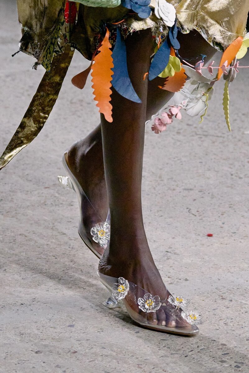 Calçado transparente decorado com flores e salto em desfile; exemplo de sapato polêmico de 2025.