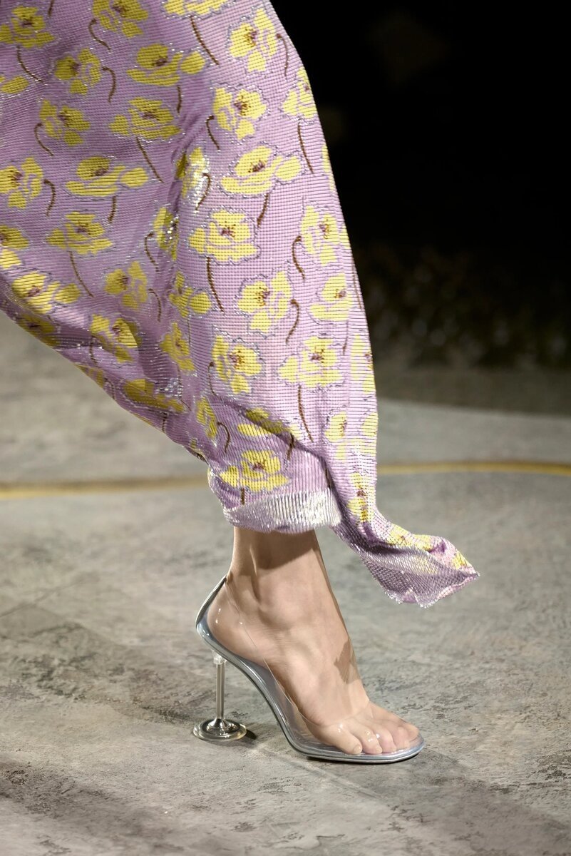 Sandália transparente com salto metálico alto, usada com vestido floral roxo e amarelo. Destaque para o design de 2025.