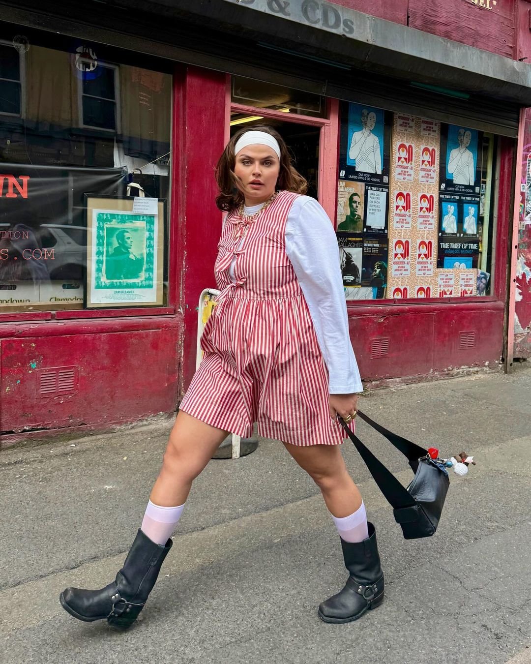 Pessoa caminhando em frente a uma loja vintage, usando sapatos de bico quadrado e vestido listrado vermelho e branco.