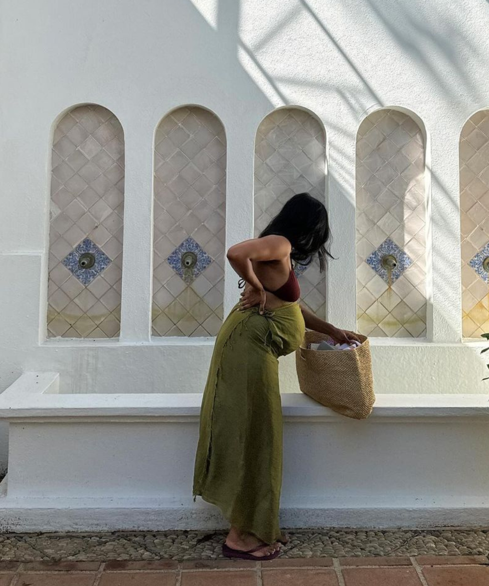 Mulher com saia verde e bolsa de palha em frente a parede decorada. Não é possível descrever sapatos bonitos e confortáveis.