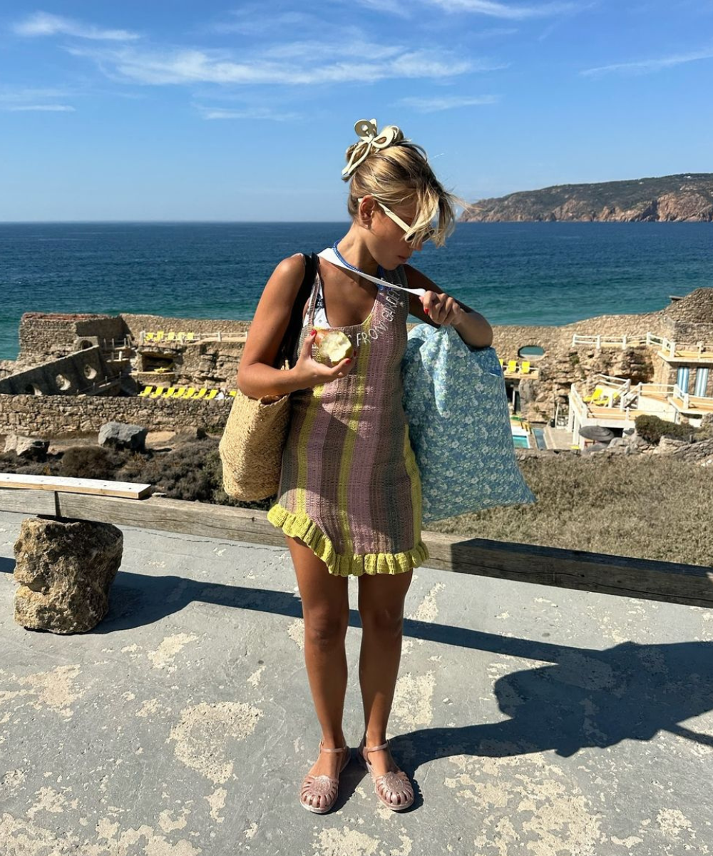 Pessoa com vestido de tricô colorido está em um cenário de praia, segurando sacolas. Usa sandálias bonitas e confortáveis.