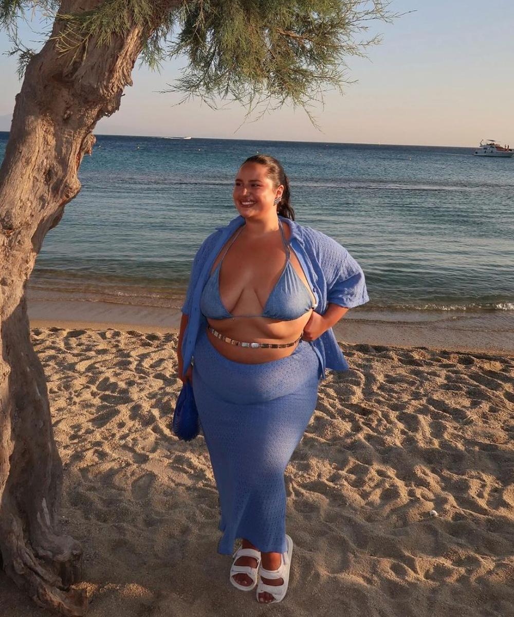 Mulher sorridente na praia ao pôr do sol, vestindo conjunto azul e sandálias brancas, sapatos bonitos e confortáveis.