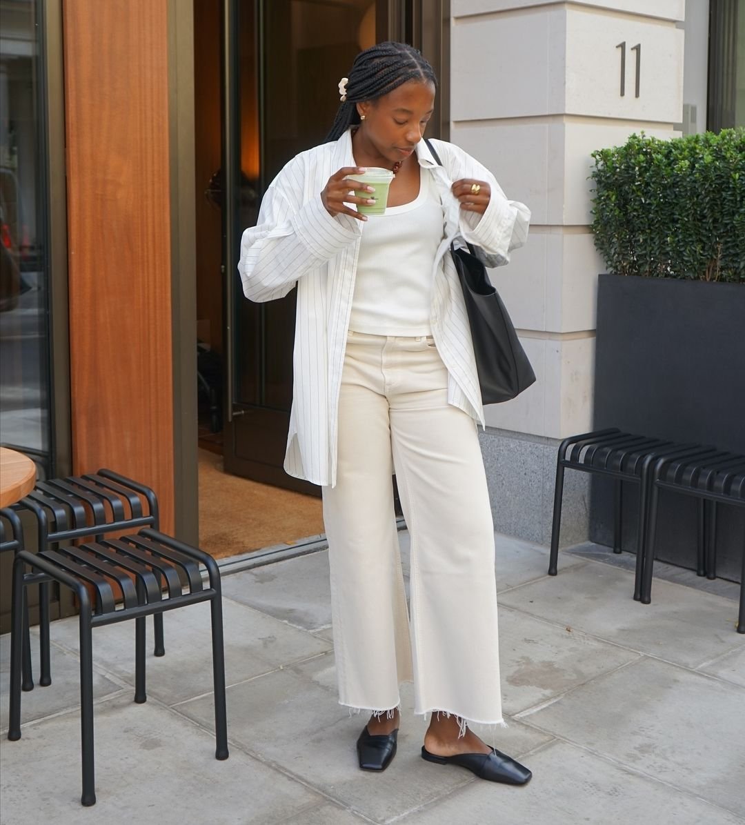 Mulher em roupas claras na calçada segura um café. Ela usa sapatos de trabalho pretos e bolsa preta.