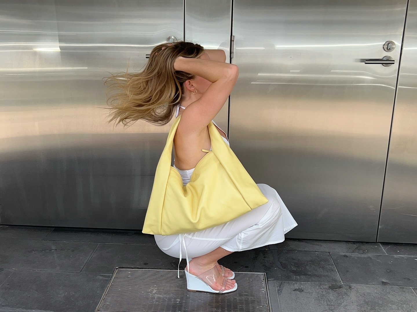 Mulher em frente a portas metálicas, com bolsa amarela e sapatos altos brancos, pose flexionada.