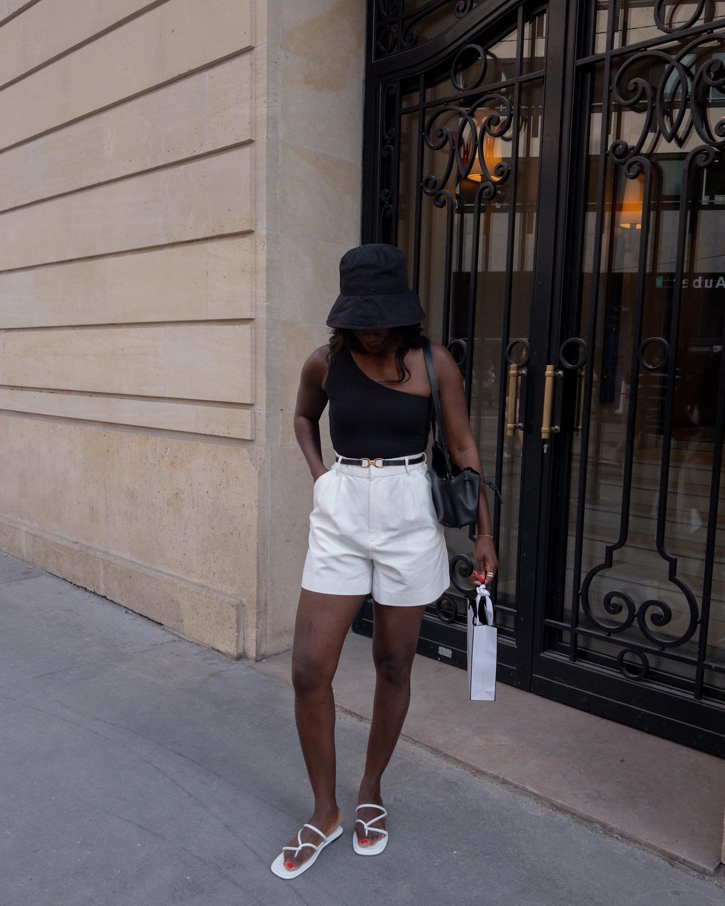 Pessoa com shorts brancos de verão, top preto e chapéu, em frente a um prédio elegante, segurando uma bolsa e um pacote.