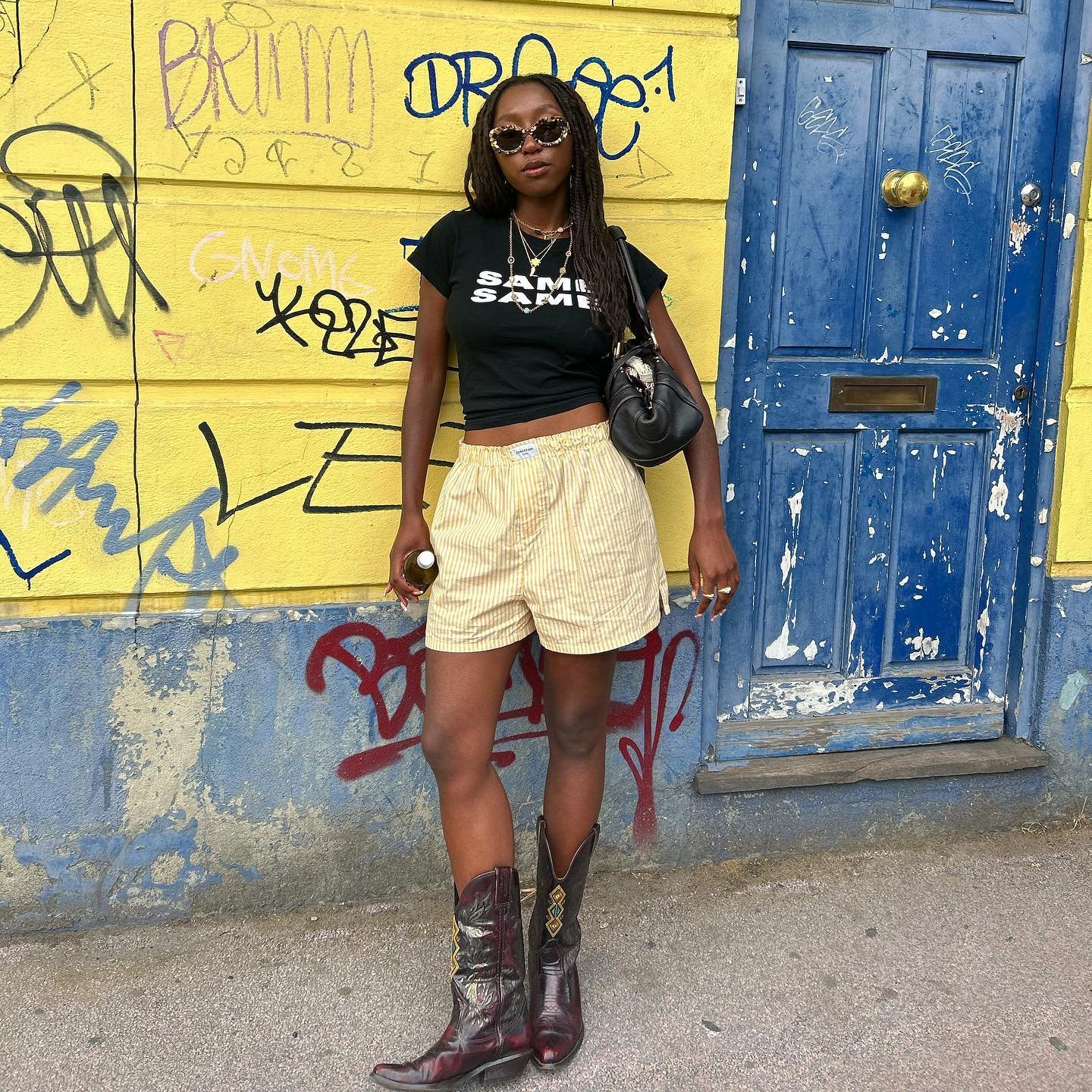 Mulher usando shorts listrados, camiseta preta e botas, posa em rua com parede grafitada e porta azul.