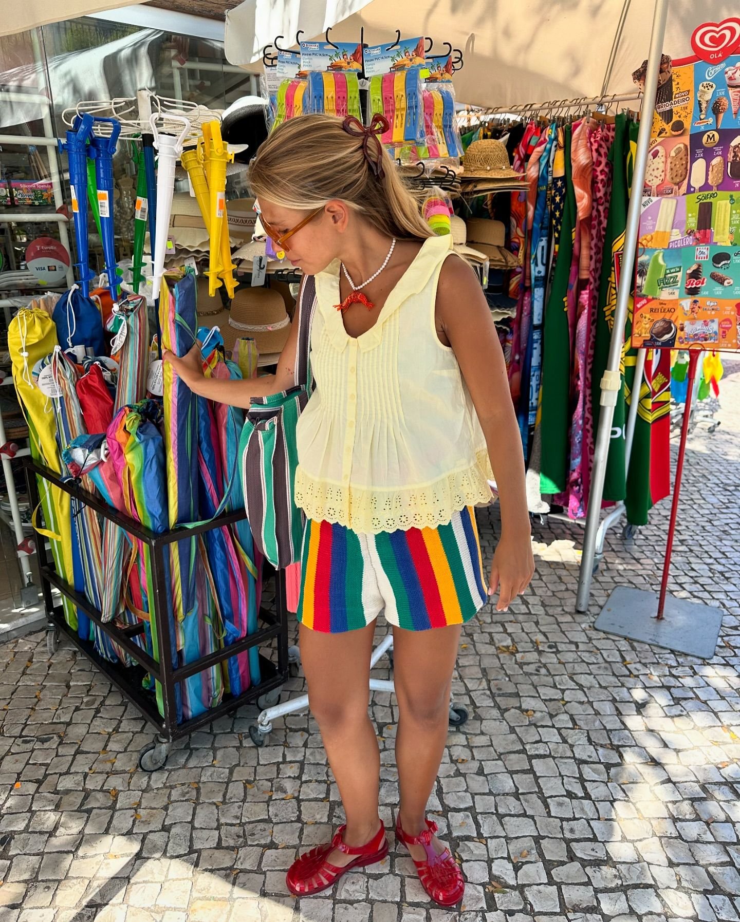 Mulher em shorts listrados coloridos explora feira ao ar livre, cercada de guarda-sóis e produtos de verão.