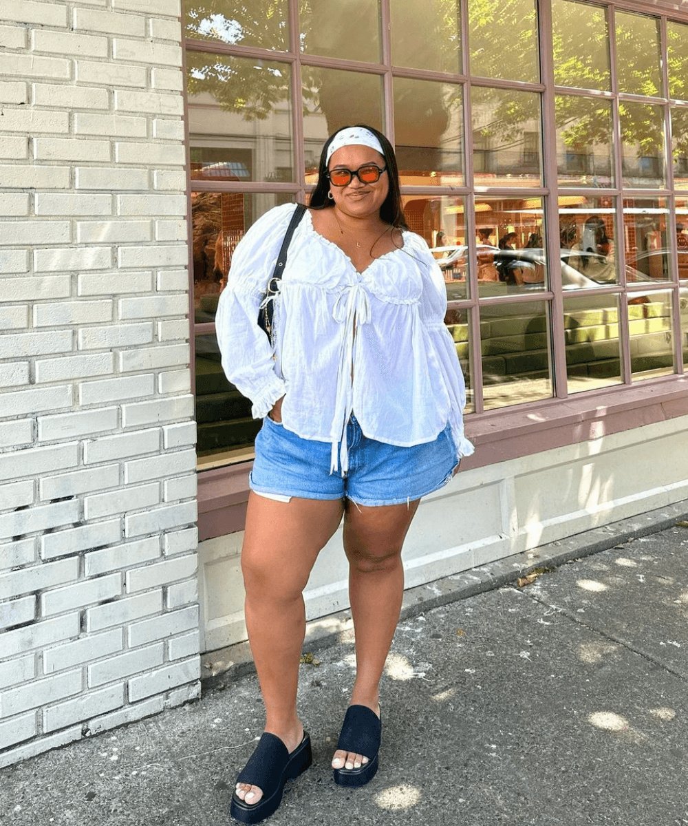 Pessoa sorrindo com blusa branca e shorts jeans para o verão, em frente a prédio com janelas.