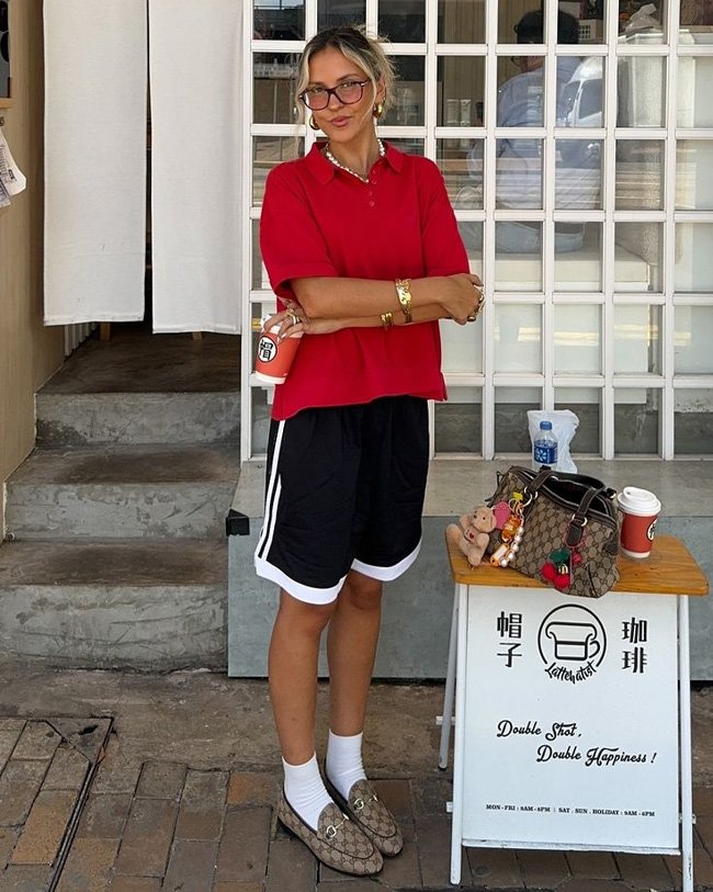 Pessoa com blusa vermelha e shorts pretos, estilo casual para o verão, em frente a uma cafeteria.