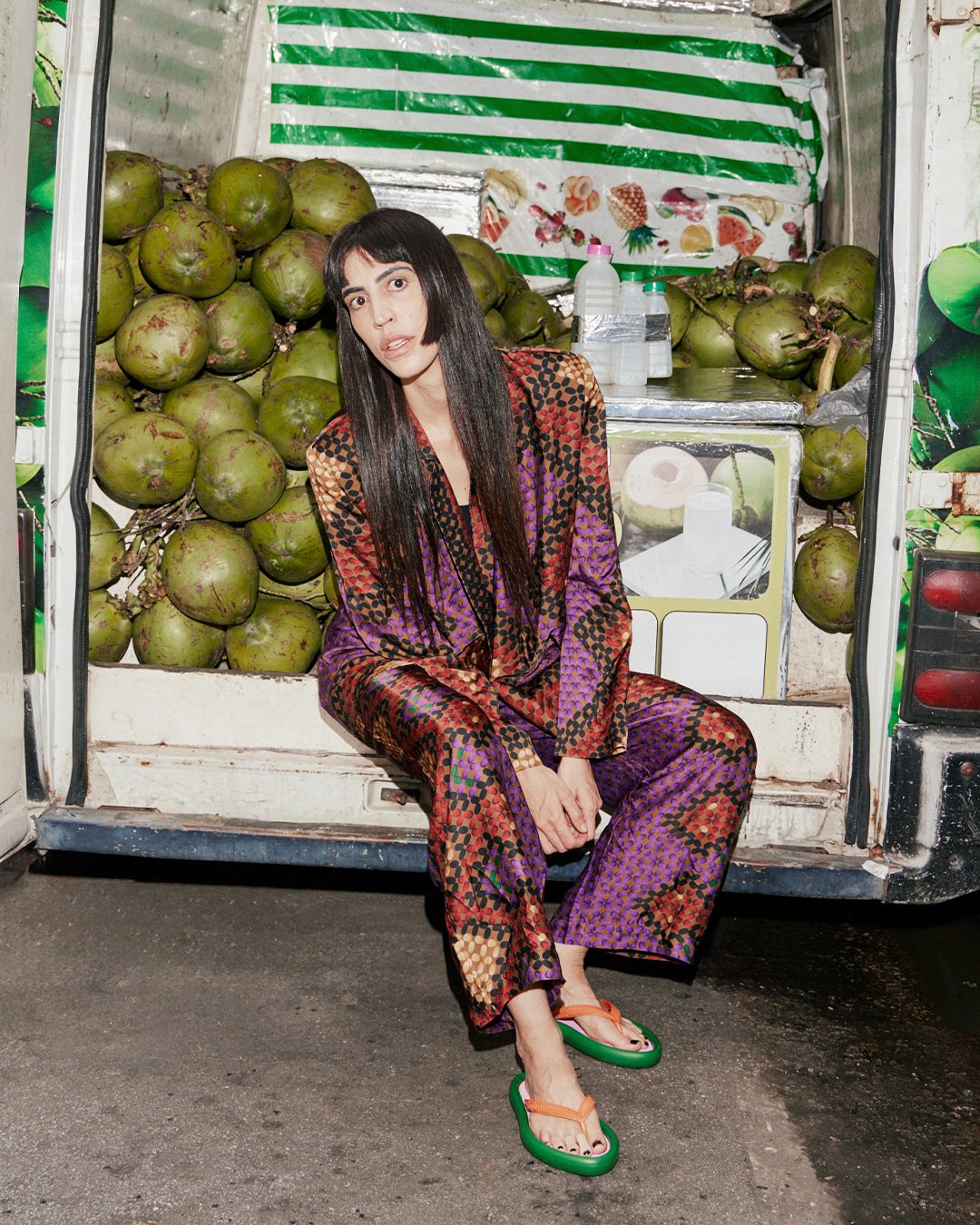 Mulher com roupa colorida sentada em caminhão com cocos, usando chinelo no verão.