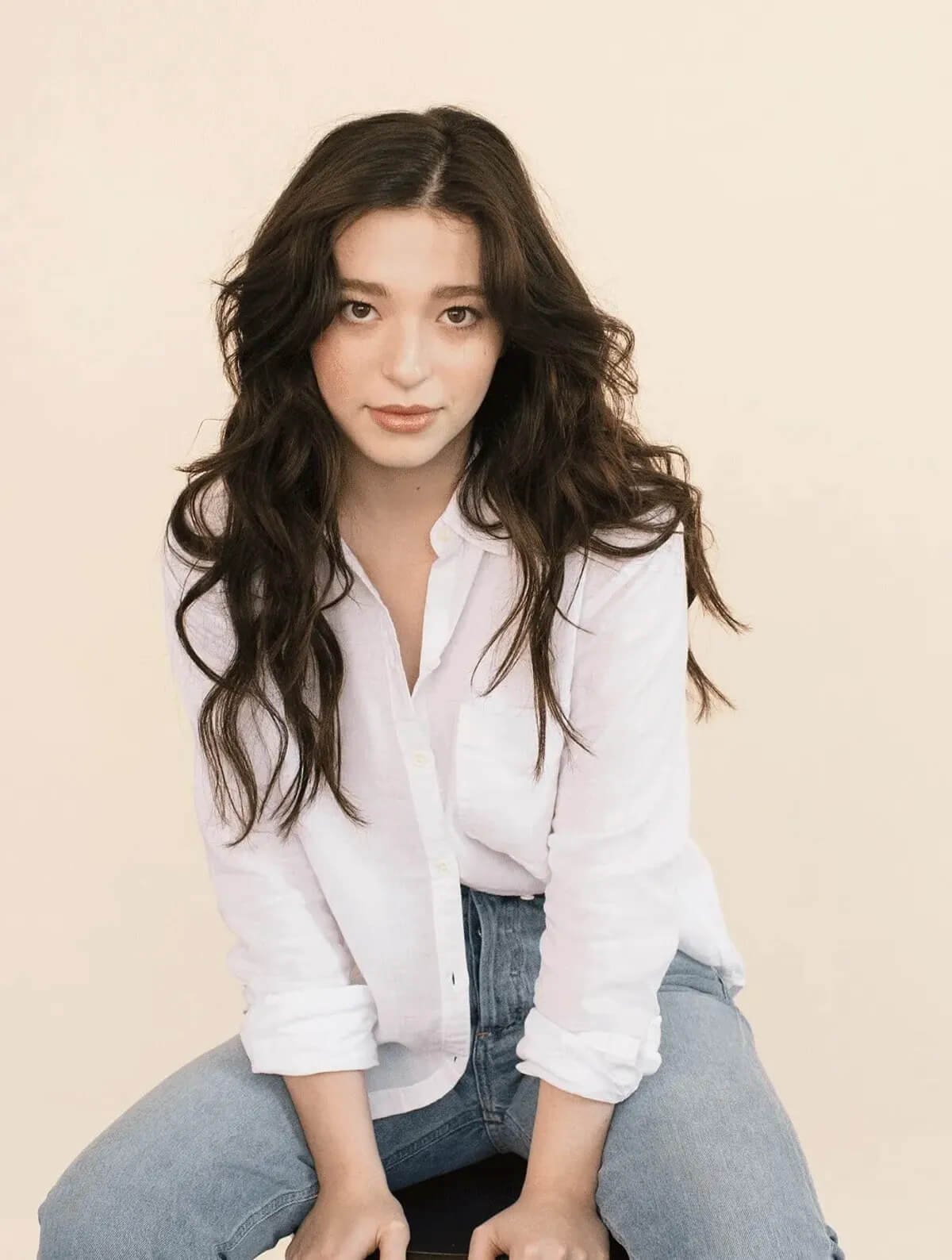 Mikey Madison com camisa branca e cabelo ondulado sentada, olhando diretamente para a câmera.