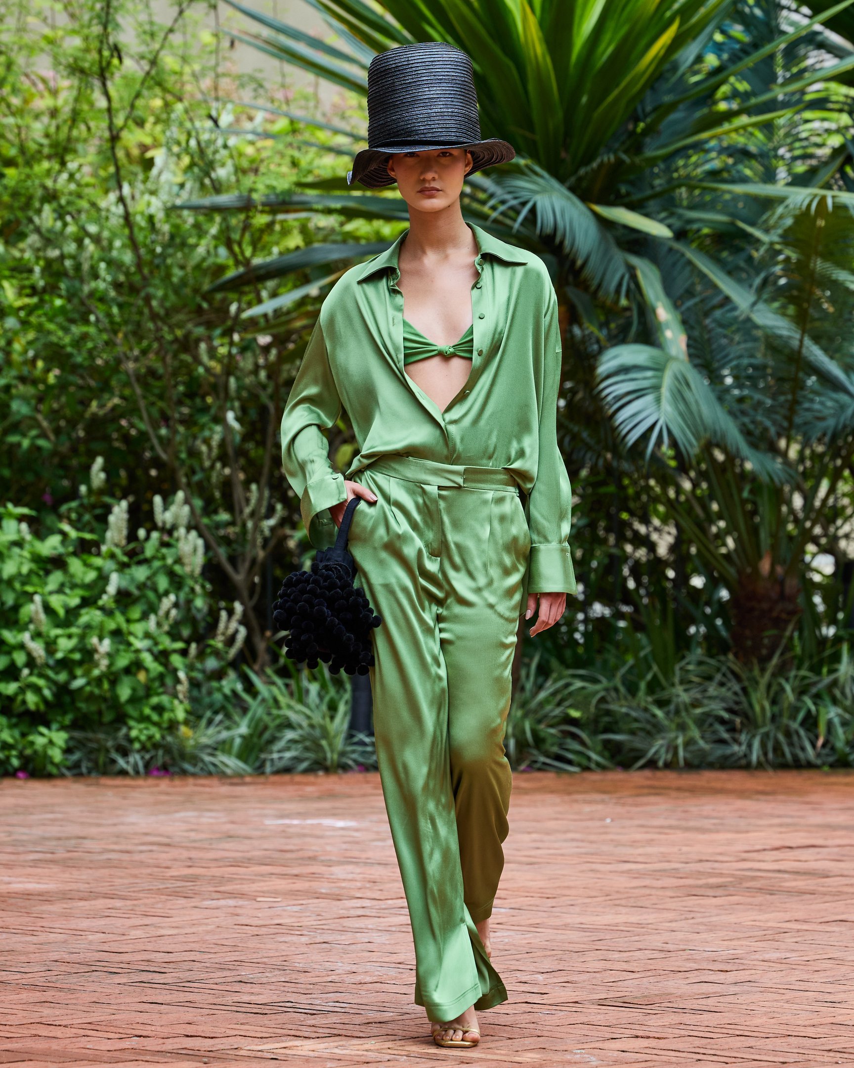 Modelo desfila com traje verde elegante, combinando camisa aberta e calça fluida, junto a um chapéu preto estiloso.