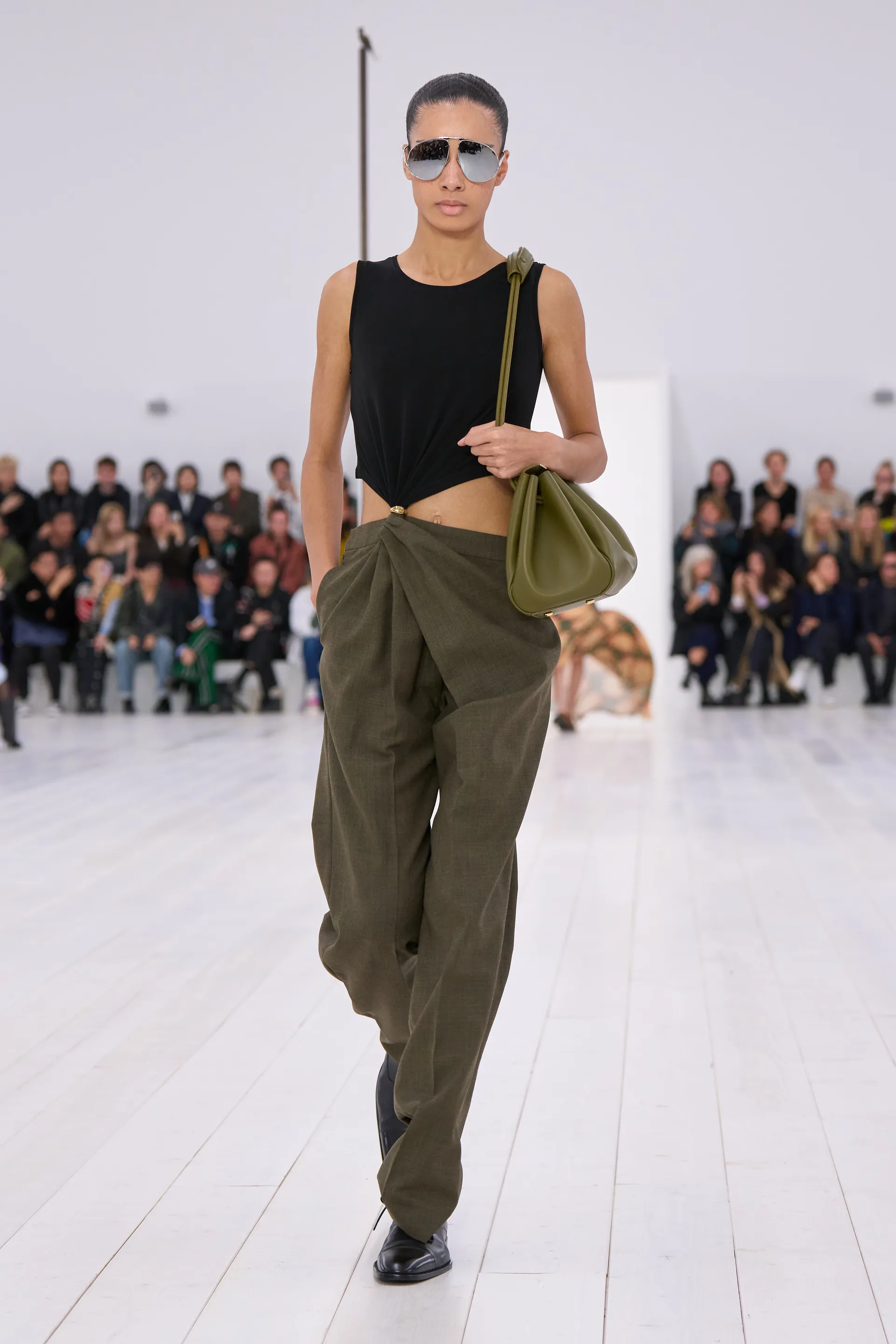 Modelo usando calça larga verde-oliva e blusa preta em desfile, destacando cores do verão 2025.