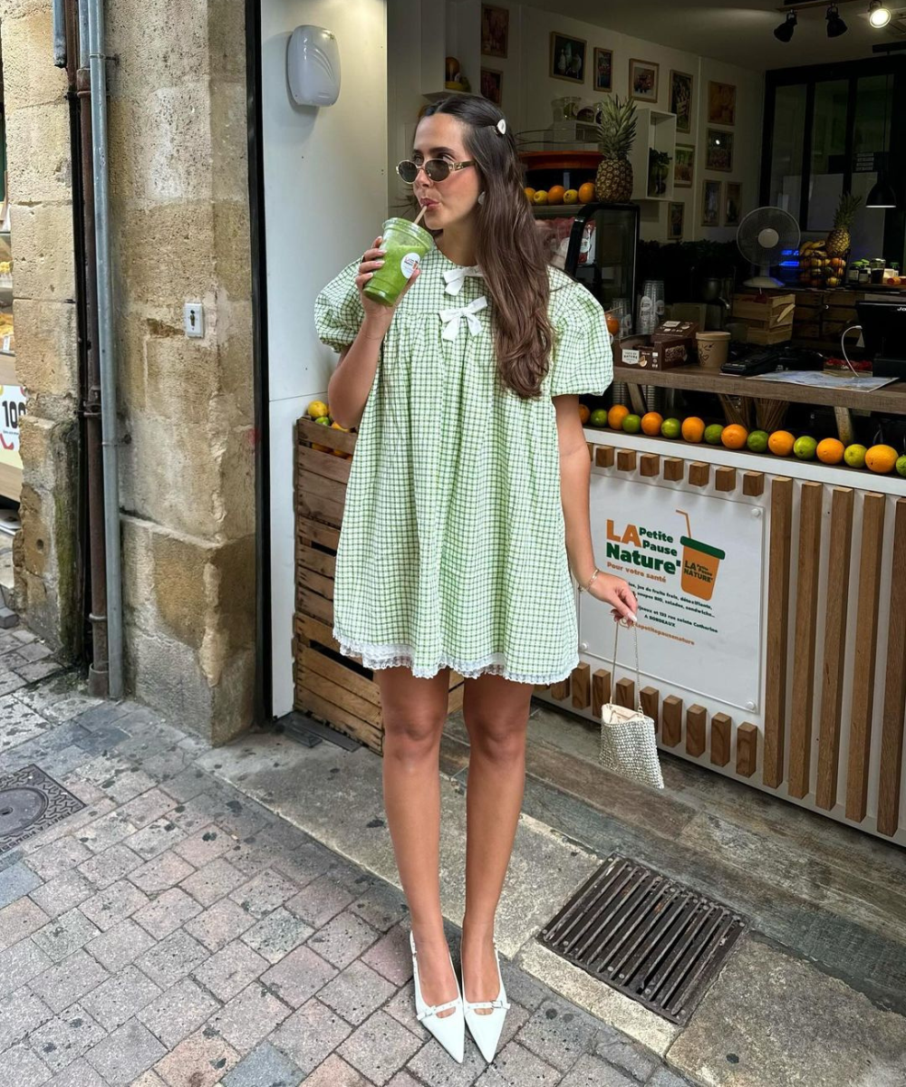 Mulher em vestido xadrez verde, tomando suco verde com canudo em frente a uma loja de frutas. Um dos vestidos soltinhos que servem muito bem para os dias quentes.