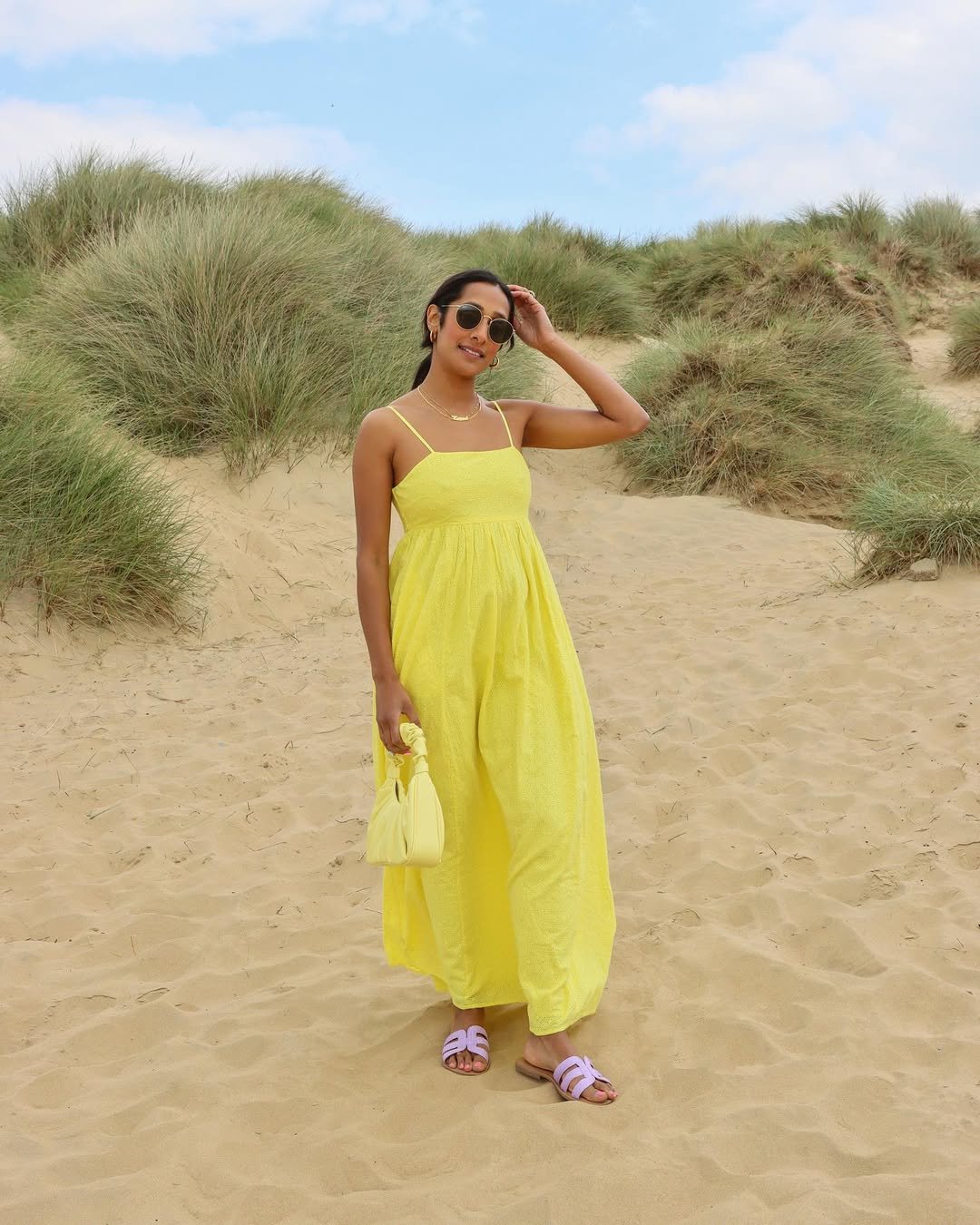 Mulher de vestido amarelo e bolsa colorida está em uma praia com vegetação ao fundo.