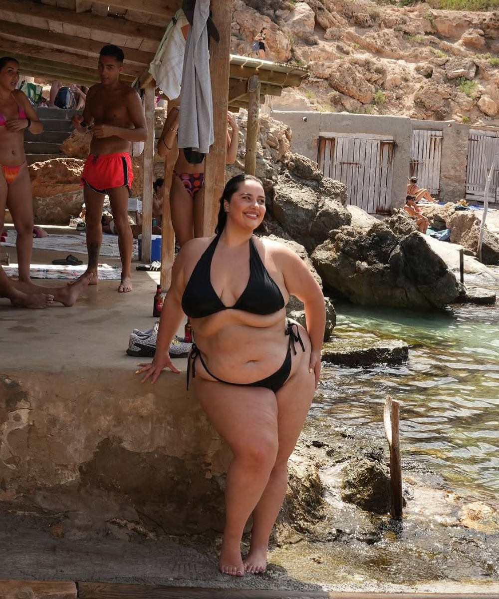 Mulher de biquíni preto em cenário de praia rochosa, transmitindo alegria e conforto.