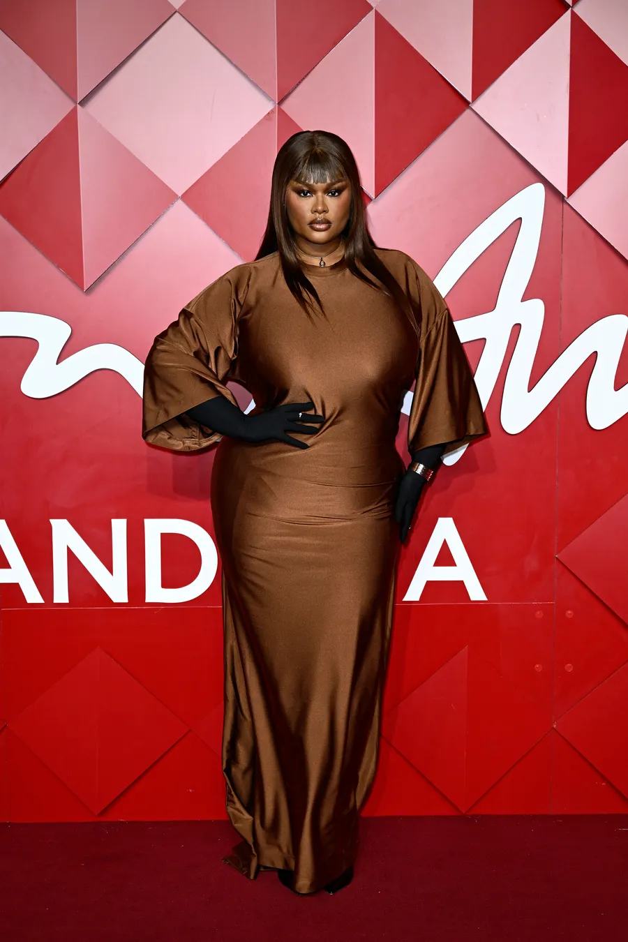 Mulher em vestido marrom com luvas pretas, em frente a um fundo vermelho no British Fashion Awards 2024.