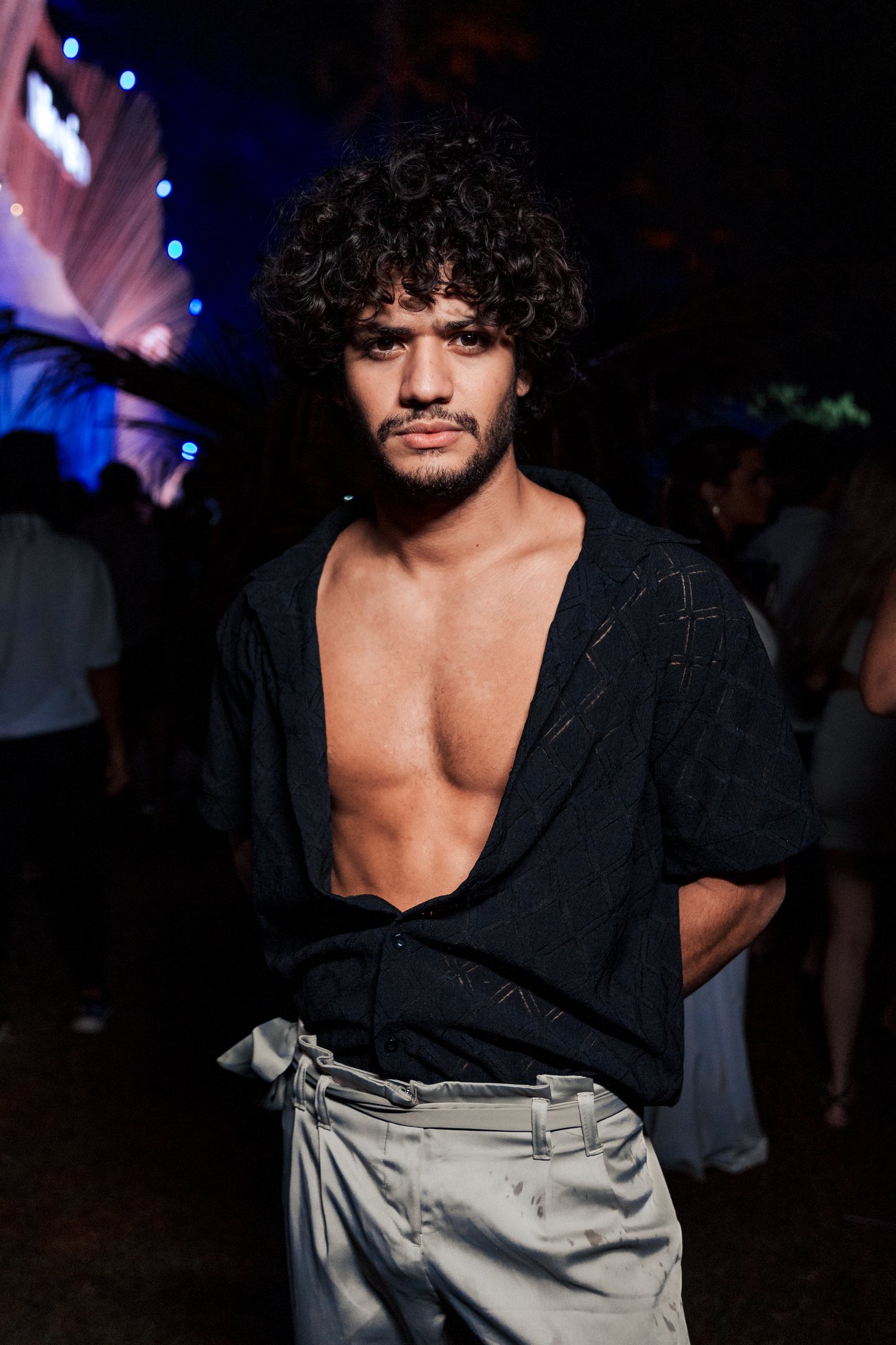 Homem de cabelo cacheado em festa à noite, uso de camisa aberta. Réveillon Amoré ao fundo.