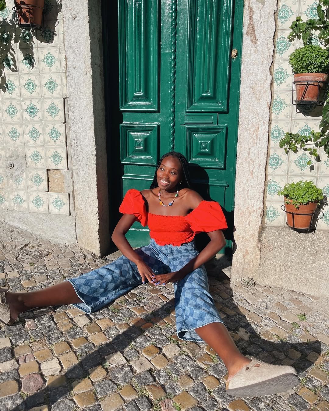 Pessoa sentada em frente a uma porta verde com blusa vermelha, calça jeans estampada e colar colorido.