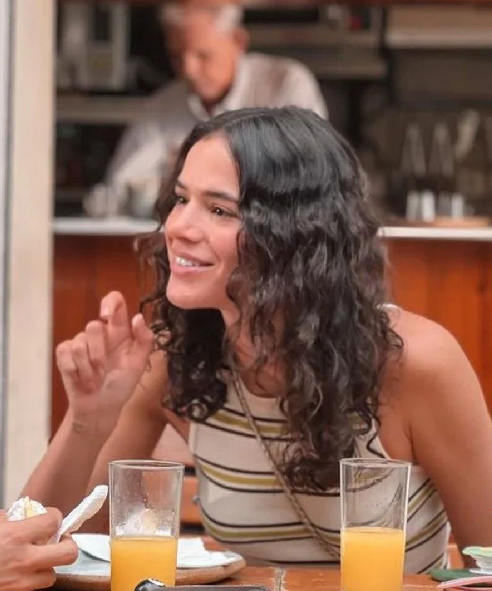 Pessoa sorrindo com cabelo cacheado, sentada em uma mesa com sucos de laranja à frente. cabelo da bruna marquezine