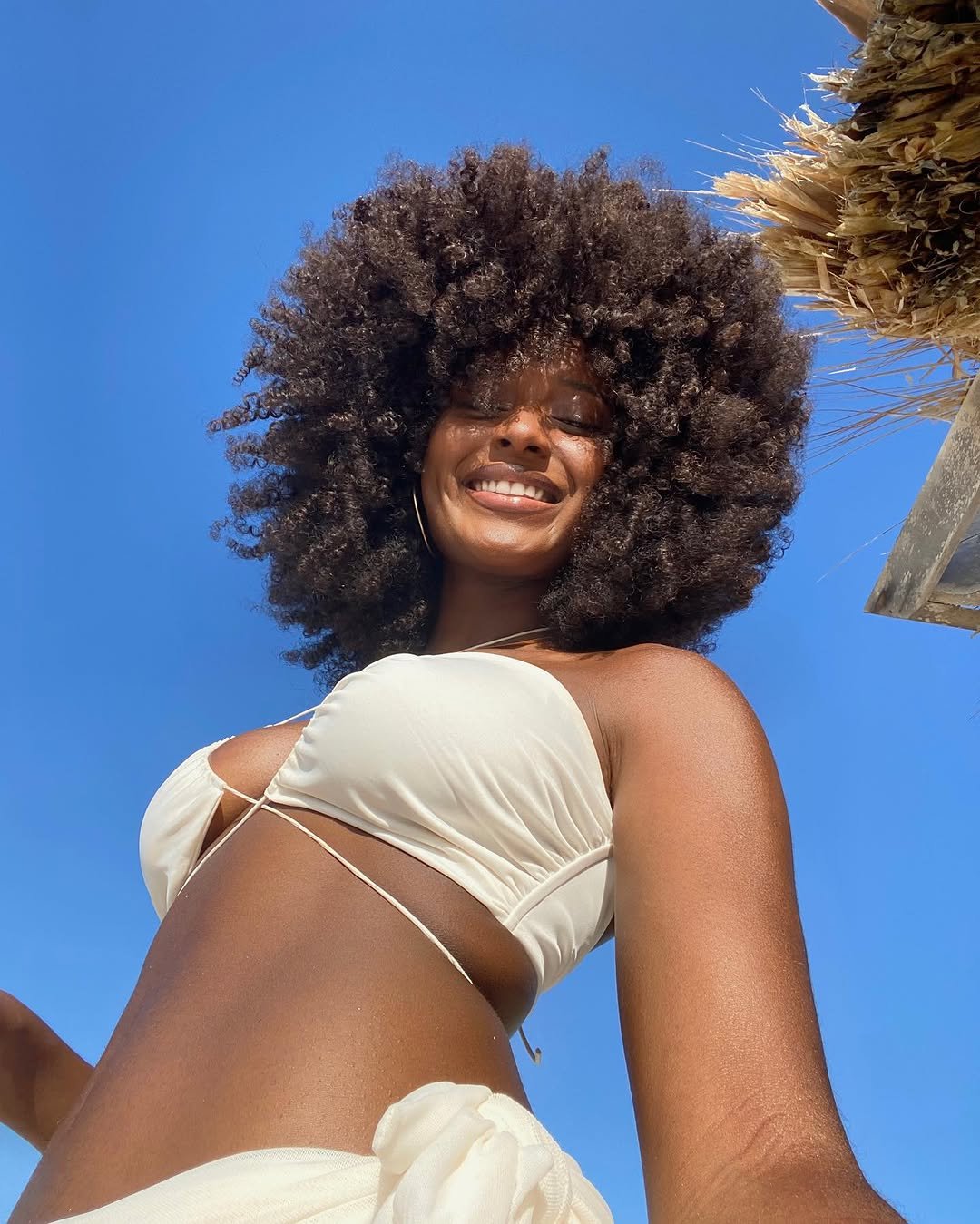 Mulher sorridente com cabelo cacheado e biquíni branco em ambiente ensolarado, lembrando um nécessaire de praia.