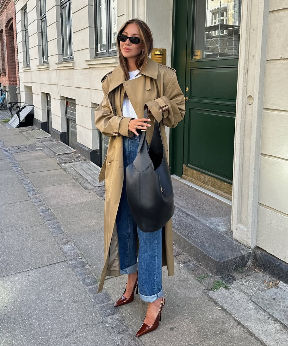 Mulher de óculos escuros veste casaco bege, jeans e camiseta básica branca em calçada urbana. Ideias de look com camisetas básicas.