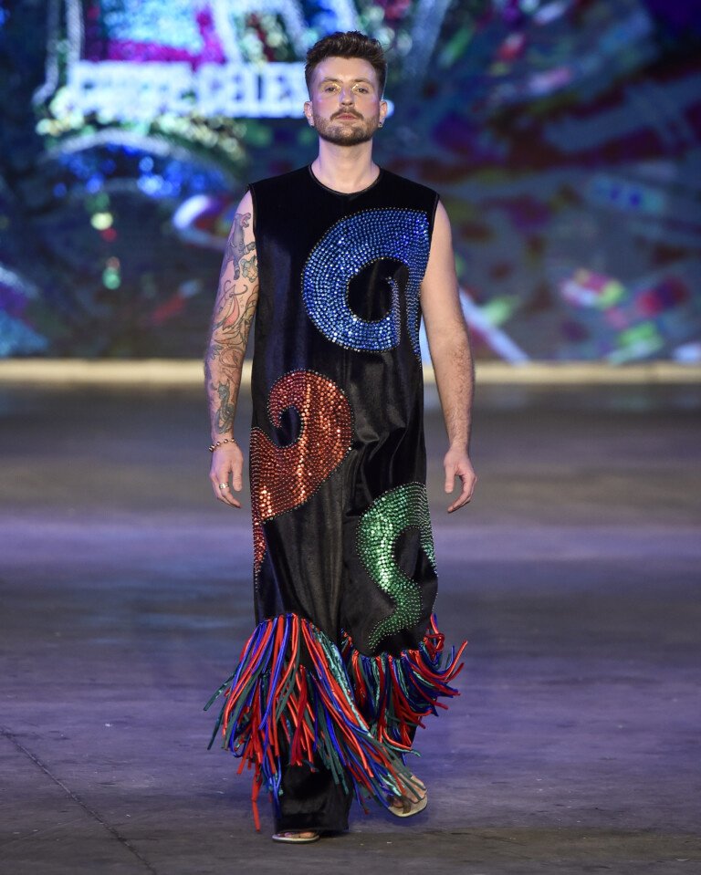 Desfile de moda com traje preto decorado com lantejoulas coloridas e franjas, em evento do tipo Casa de Criadores.