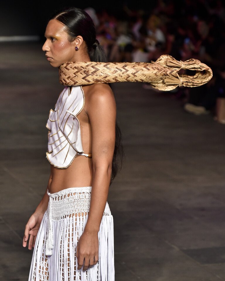 Modelo desfilando com roupa tribal e acessório de cobra em um evento de moda.