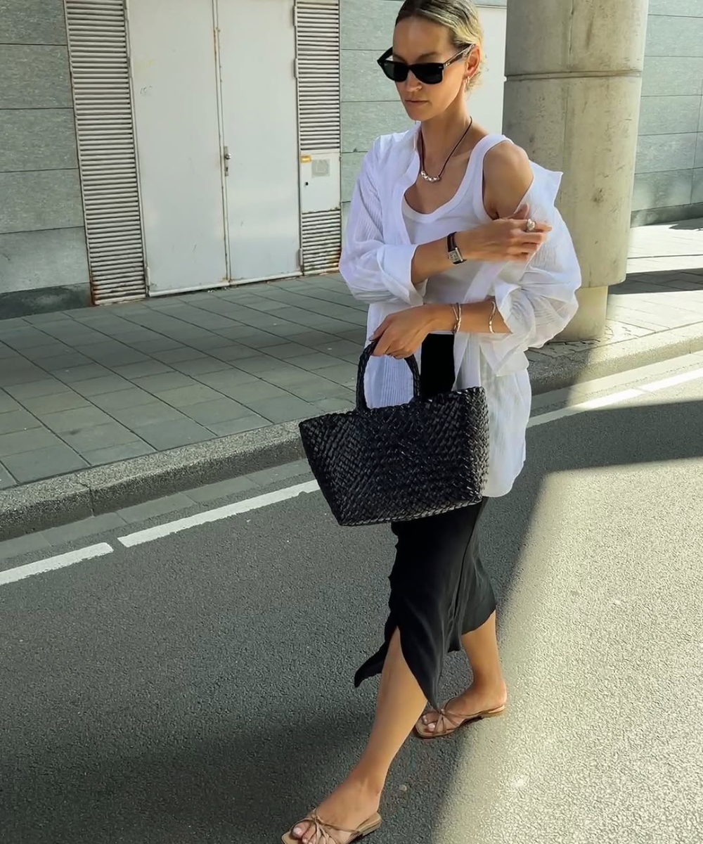 Mulher caminhando na rua com bolsa preta trançada, vestido preto e camisa clara.