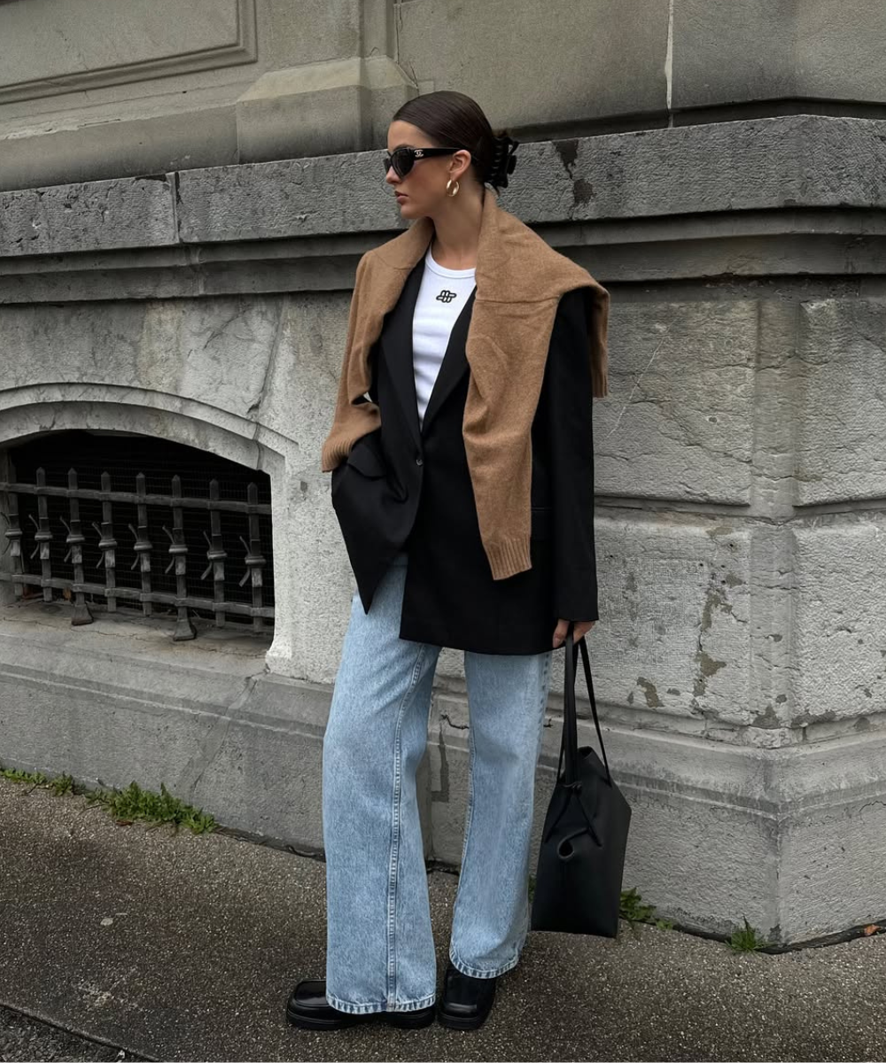 Mulher elegante veste blazer preto, calça jeans larga e bolsa preta, com suéter marrom nos ombros.