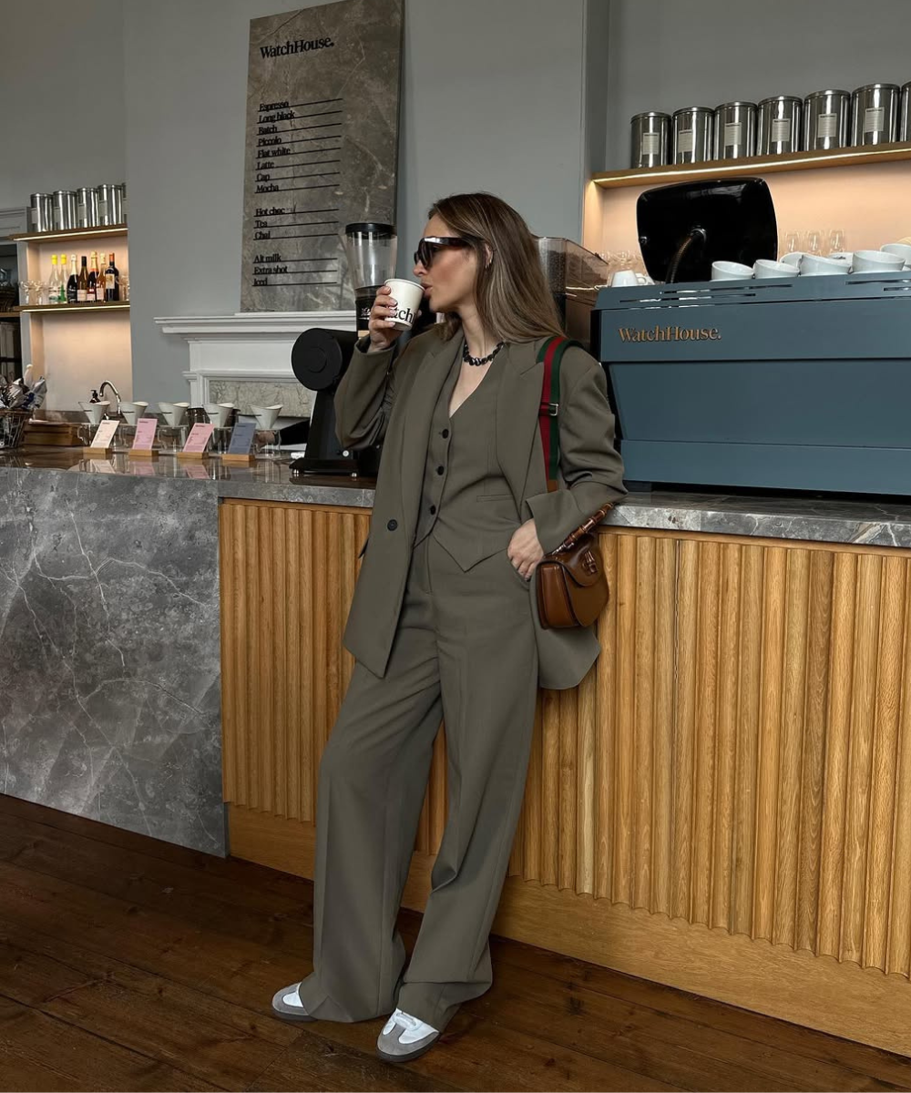 Mulher com look monocromático bege, óculos escuros e bolsa, tomando café em uma cafeteria elegante.