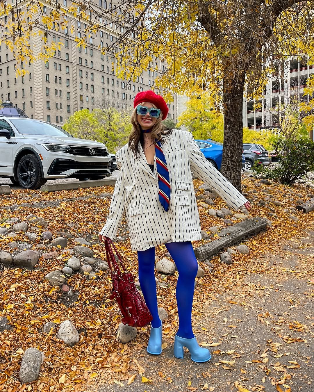 Mulher sorrindo em meio às folhas de outono, vestindo blazer listrado e acessórios vermelhos e azuis. Cor de cada signo