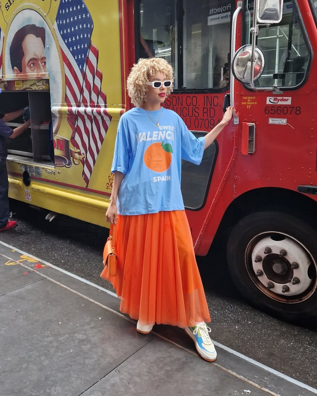 Pessoa com cabelo cacheado loiro usa camiseta azul e saia laranja em frente a caminhão vermelho. Cor de cada signo