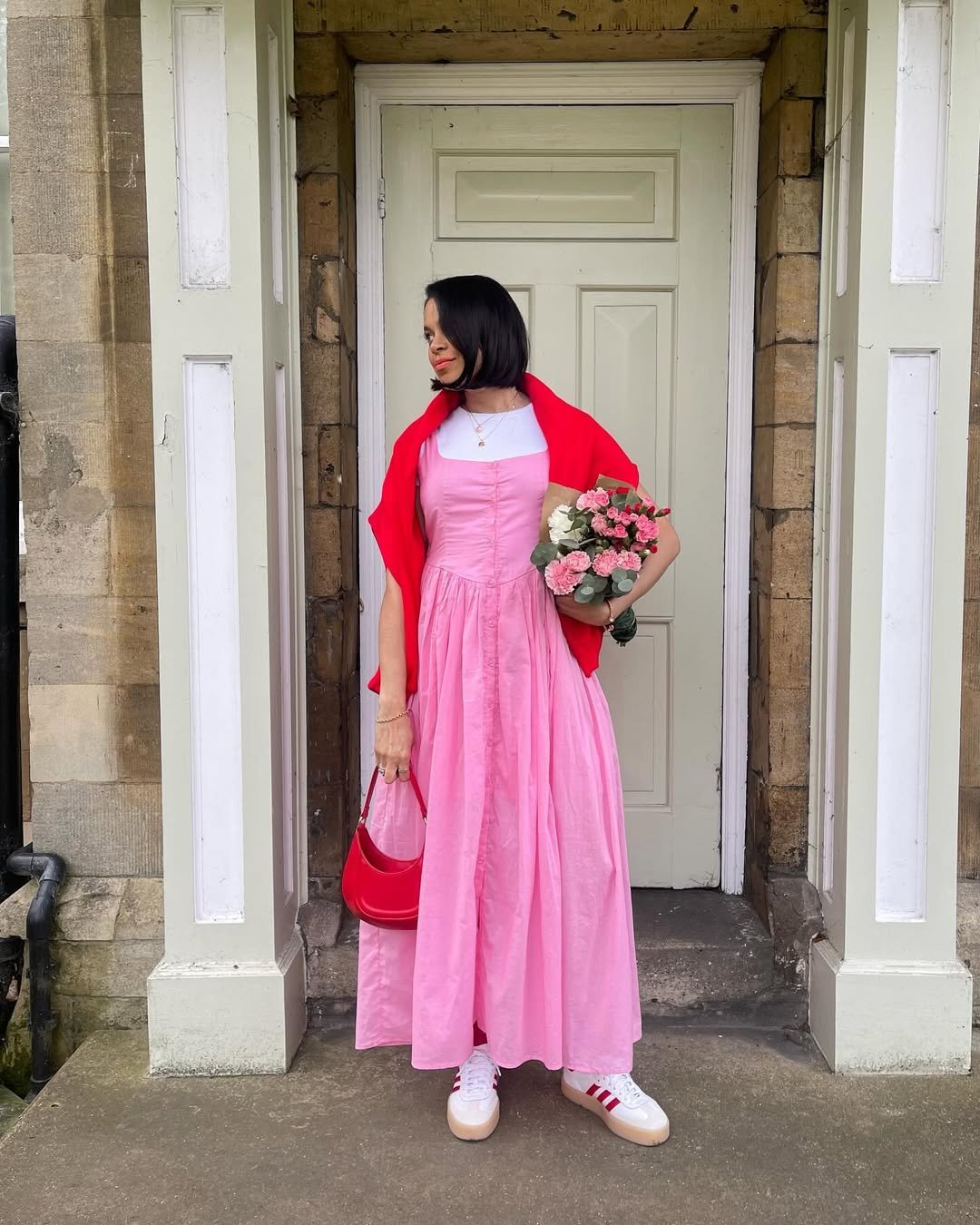 Mulher com vestido rosa, casaco vermelho e tênis, segura buquê de flores na frente de porta branca. Cor de cada signo