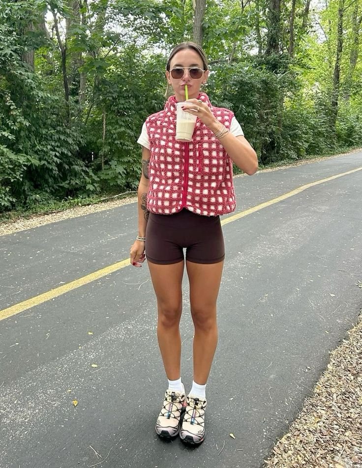 Jovem com colete vermelho, shorts e tênis em trilha arborizada, tomando bebida. Estilo descontraído e esportivo.