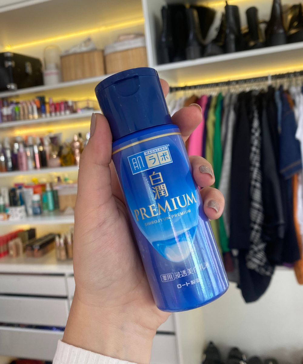 Mão segurando frasco azul de loção para a pele em um quarto com estante ao fundo. Porduto que mostra ser um 'dupe' do produto Glazing Milk da Hailey Bieber.