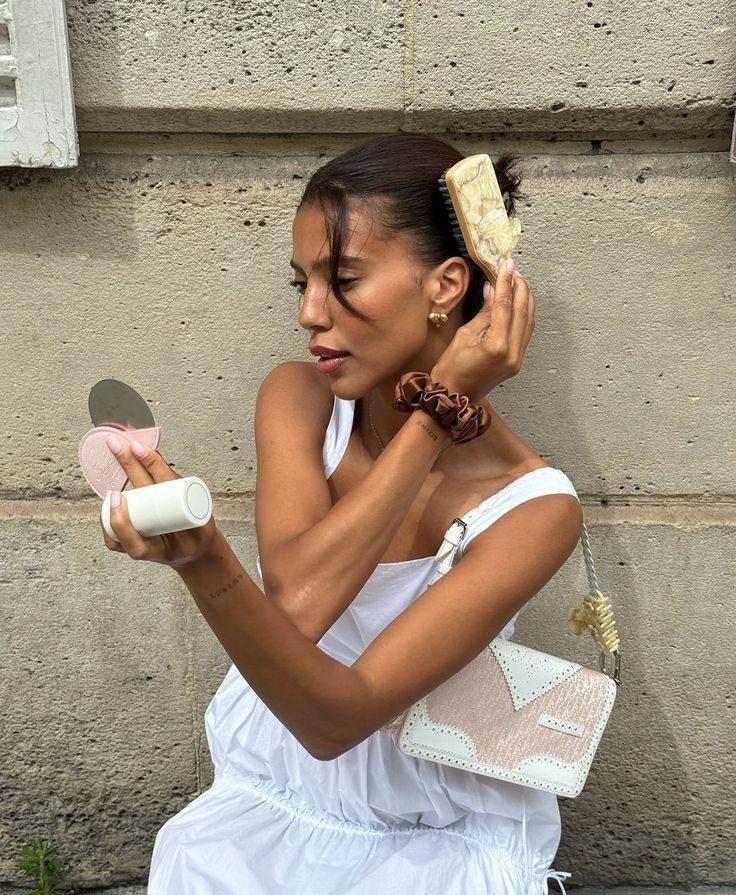 Mulher ajustando cabelo com escova, segurando espelho e produto de beleza, refletindo tendências de moda e beleza de 2024.