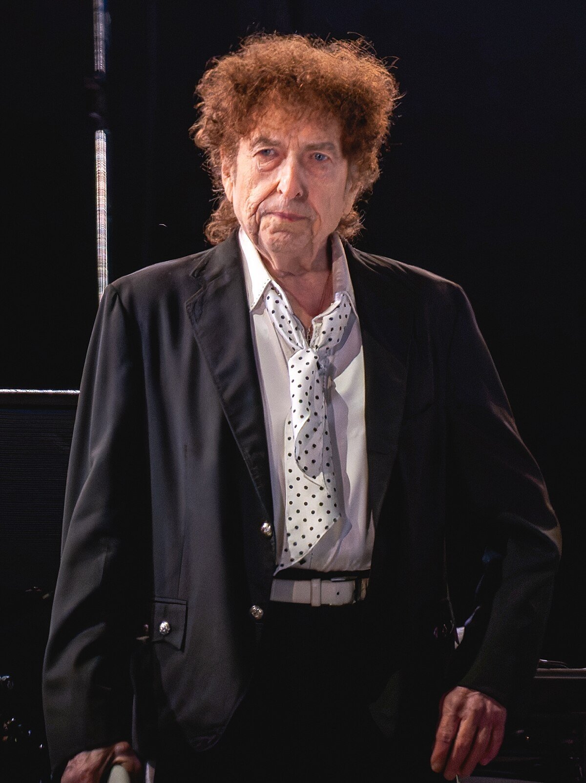 Homem de terno preto e gravata de bolinhas, em um palco, evocando um estilo de Bob Dylan.