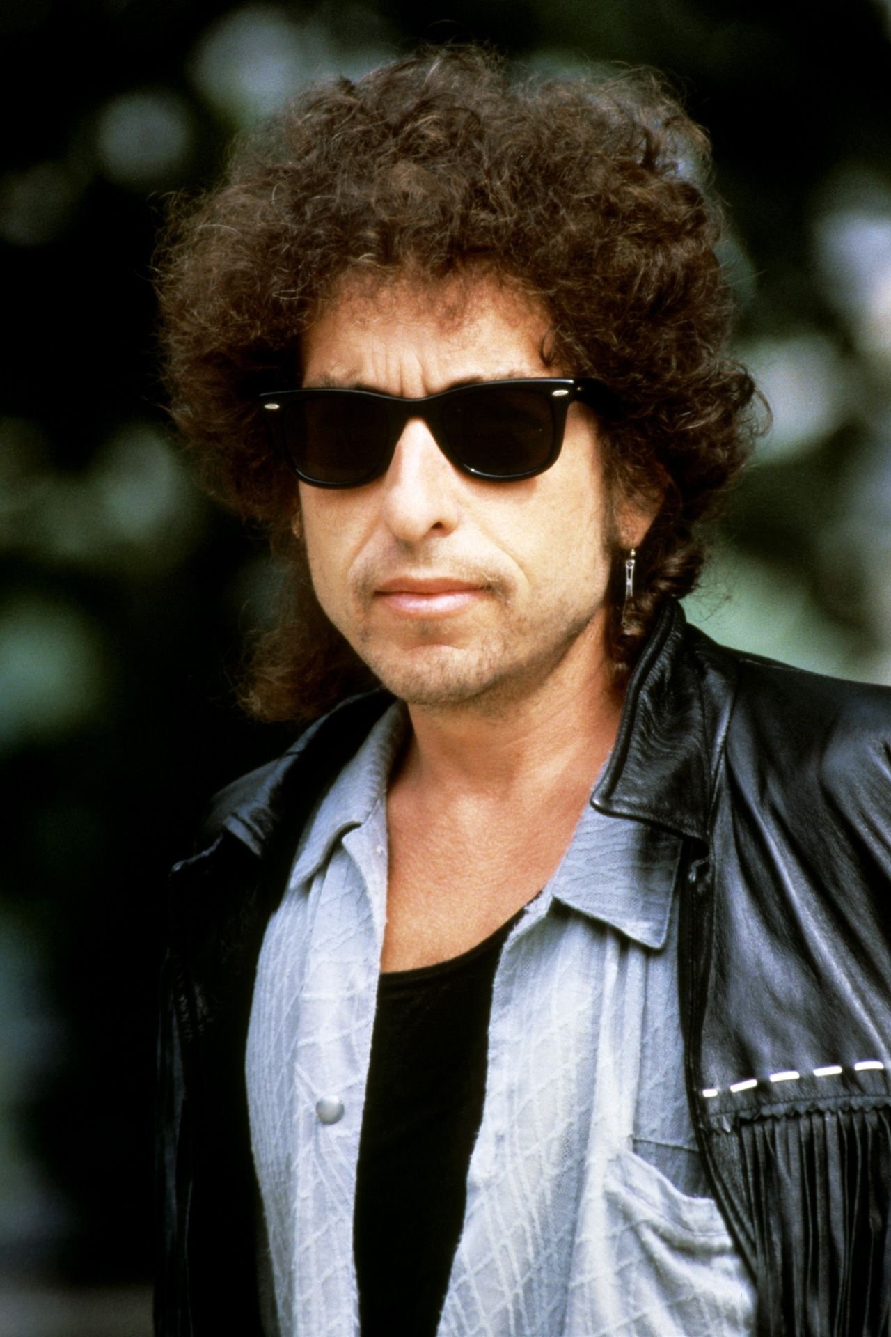 Óculos escuros e jaqueta de couro em estilo descontraído. Cabelo cacheado e expressão séria, influenciado pelo estilo de Bob Dylan.