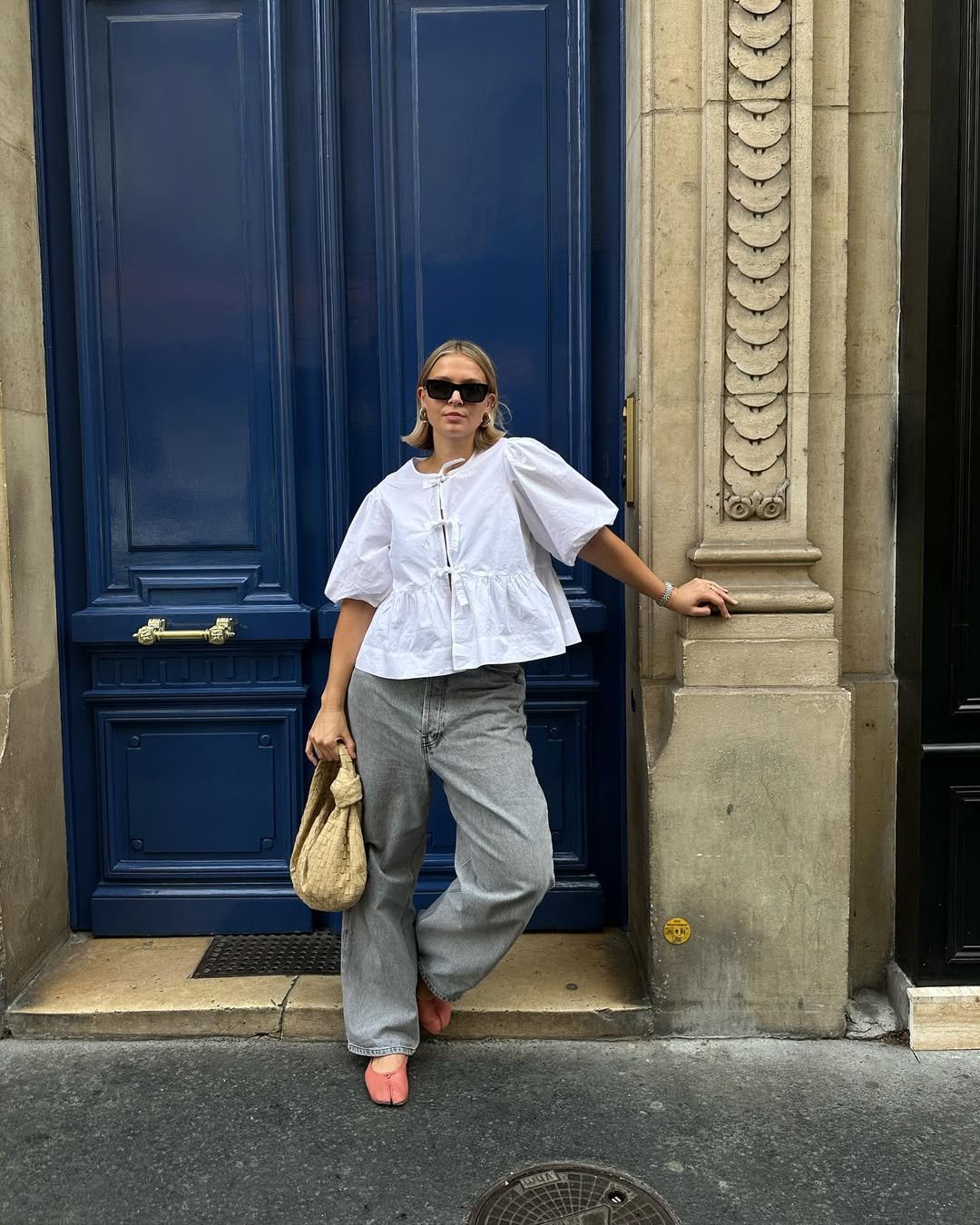 Pessoa com uma das blusas estilosas da temporada. Top branco, jeans cinza e bolsa de palha, em frente a porta azul.