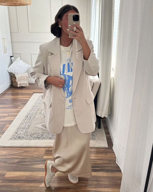 Mulher com blazer creme, camiseta gráfica, saia longa bege e tênis. Visual casual e elegante em ambiente moderno.