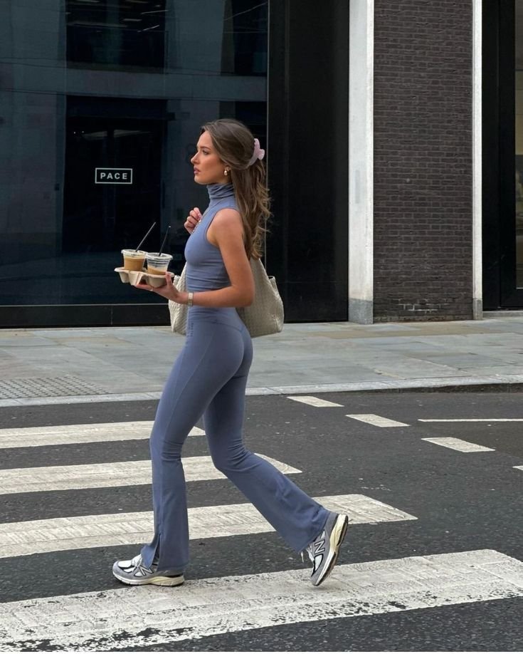 Mulher vestindo roupa fitness azul e tênis, carregando copos de café enquanto atravessa a rua.