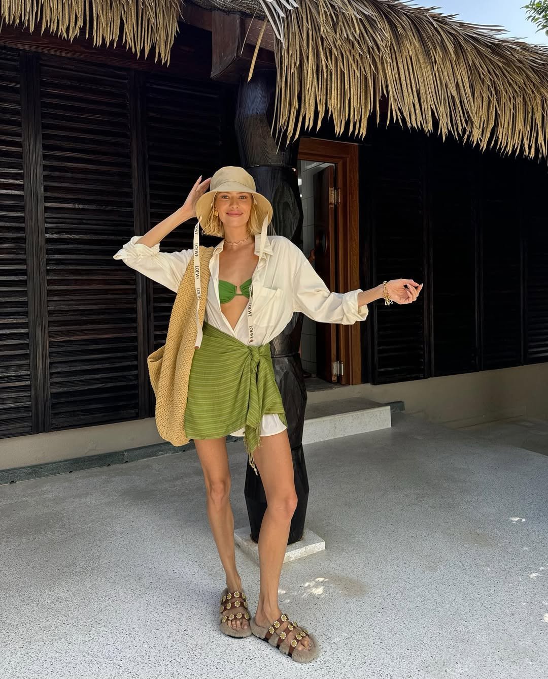 Mulher com look estiloso no verão, usando chapéu, top verde, camisa branca e sandálias com acessórios dourados.