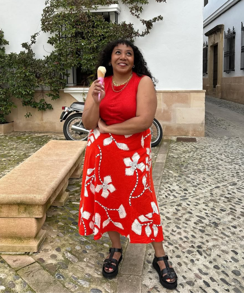 Mulher com saia vermelha florida, segurando sorvete em rua de paralelepípedo. Inspiração de looks de Natal para quem é básica.