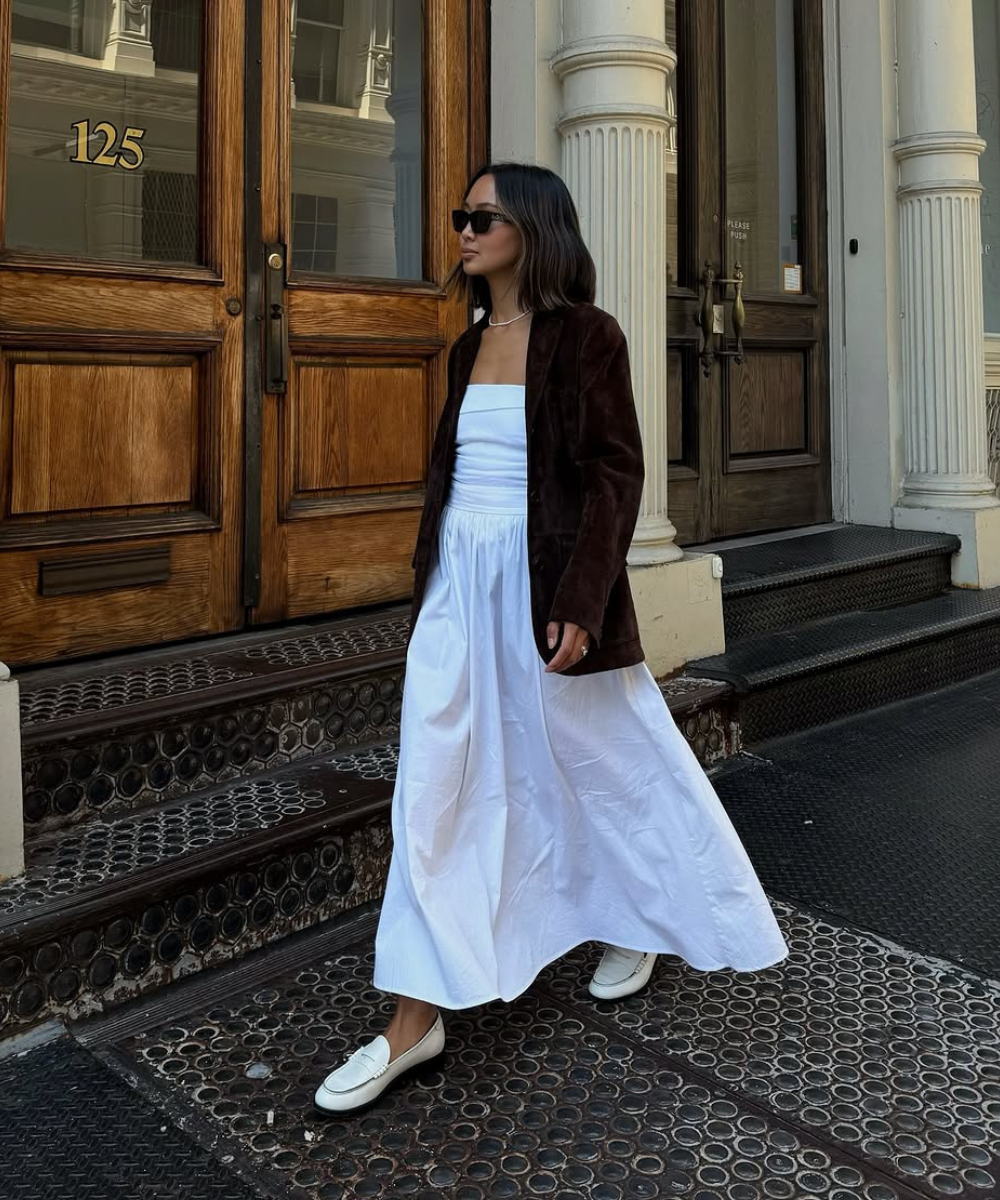 Mulher com vestido branco e jaqueta escura usa mocassim branco, em frente a portas de madeira.