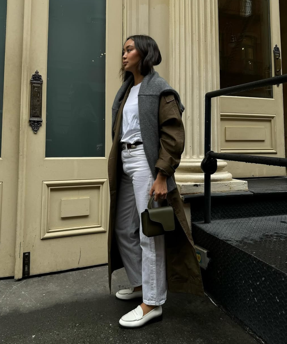 Pessoa com look casual usando mocassim branco, calça clara, camiseta branca e casaco longo verde oliva, segurando bolsa verde.