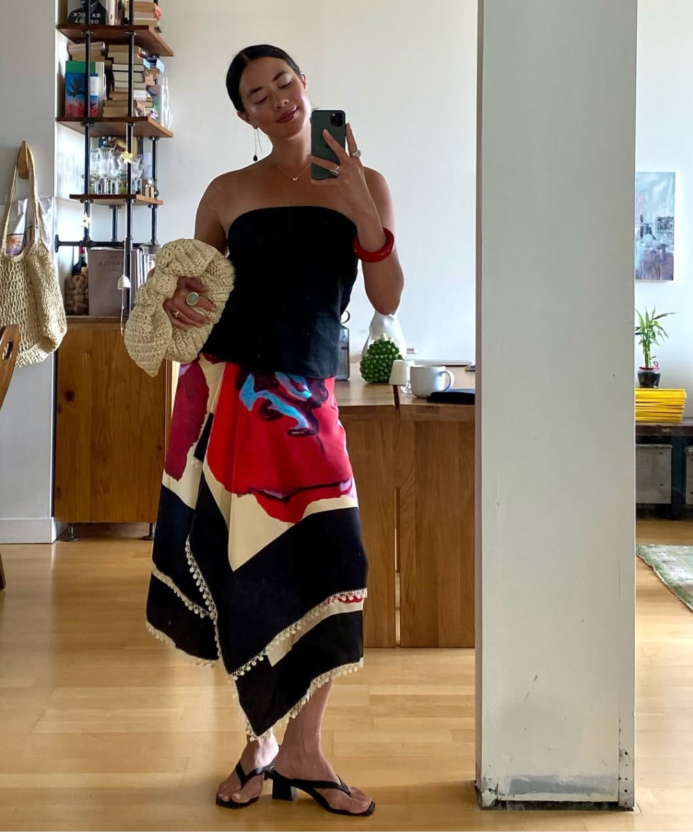 Mulher com saia estampada e blusa preta tira uma selfie. Ela carrega bolsa de palha e usa sandálias.