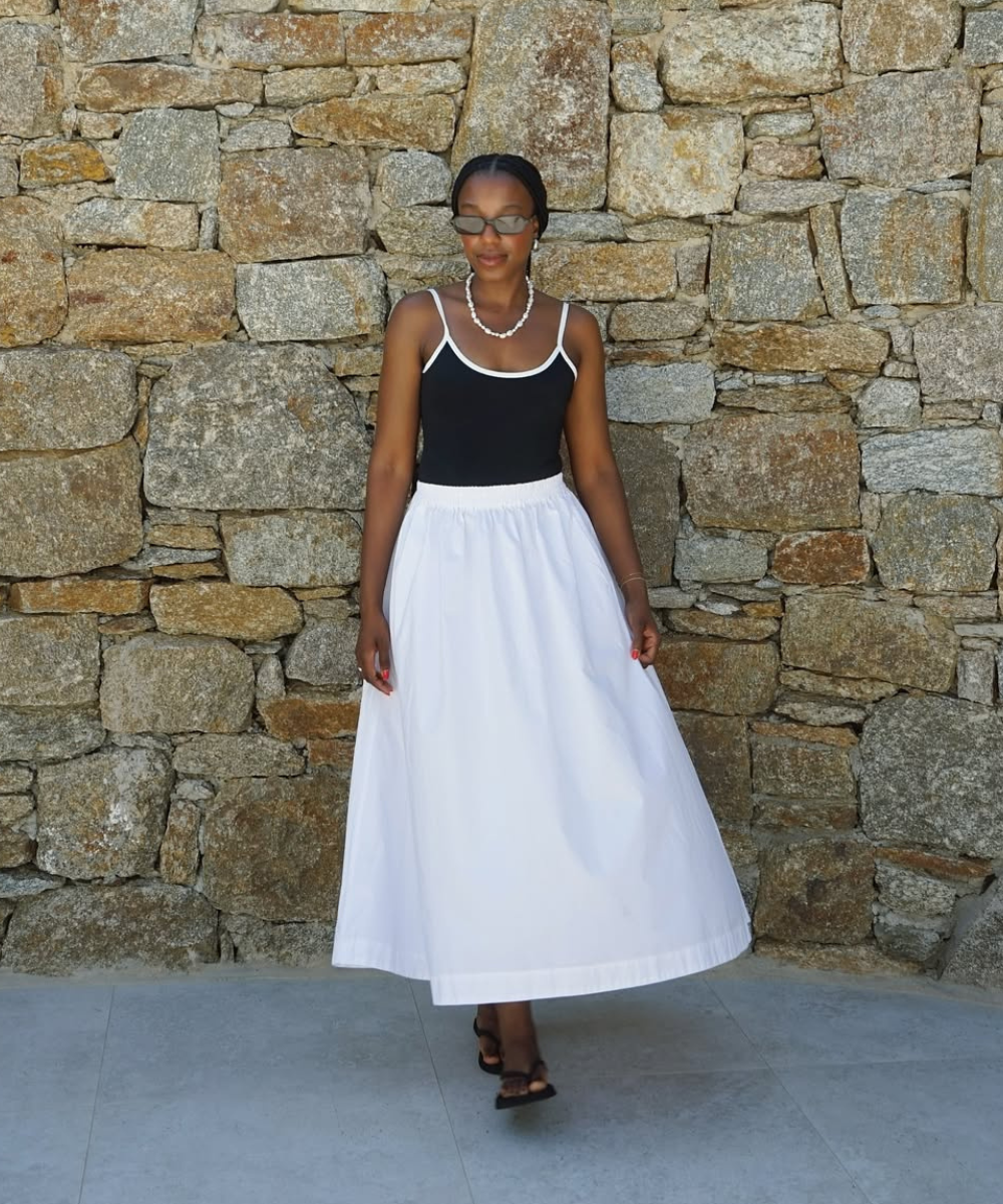 Mulher usando saia longa branca e blusa preta diante de uma parede de pedra, representando looks estilosos com saia.