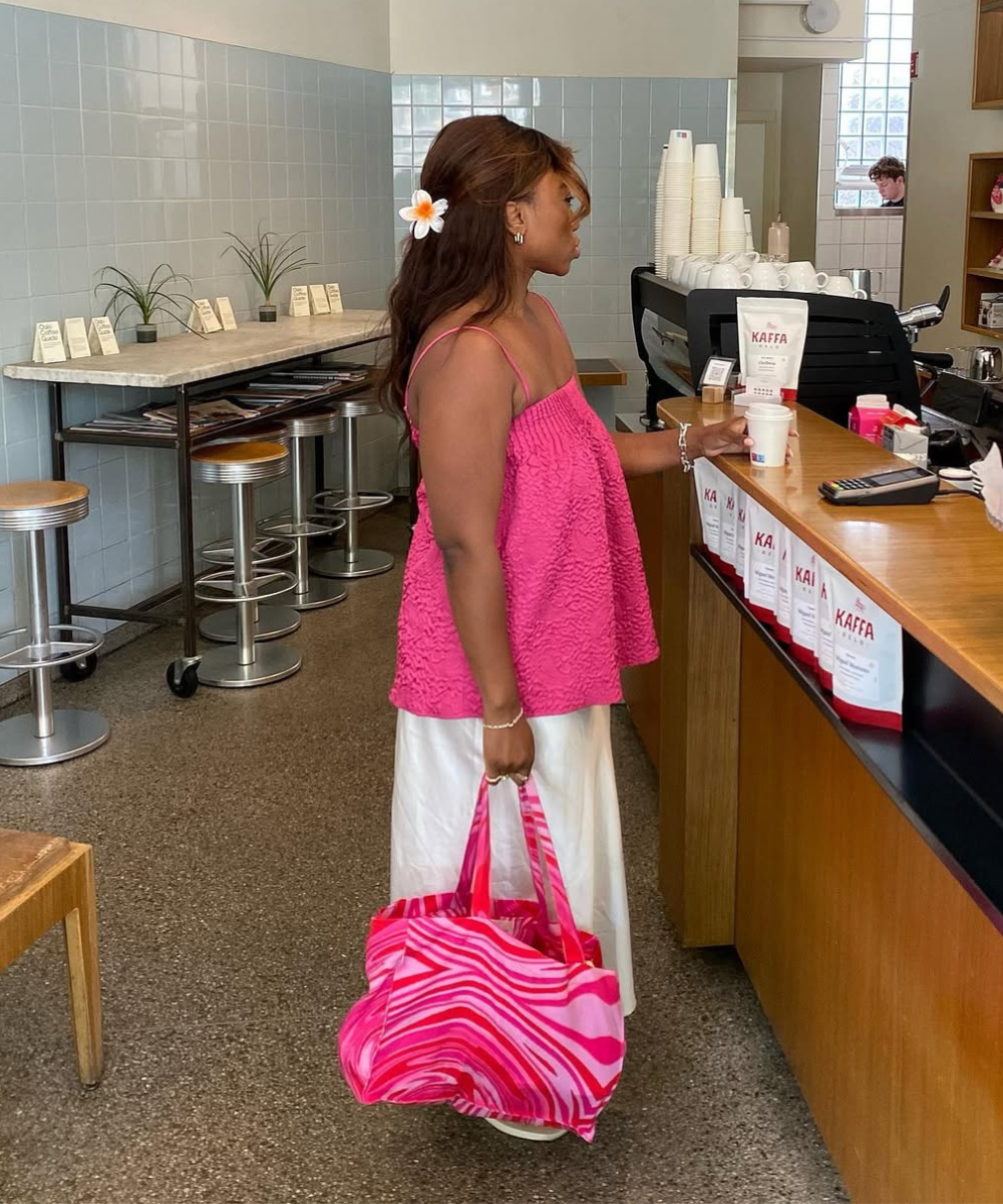 Mulher em cafeteria com regata rosa e saia branca, segurando bolsa estampada. Estilo casual e elegante.