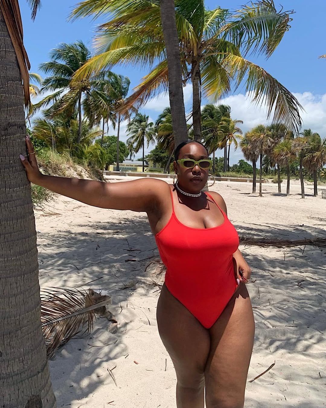 Mulher de maiô vermelho posa em praia ensolarada com palmeiras, refletindo tendências de biquíni e moda praia.